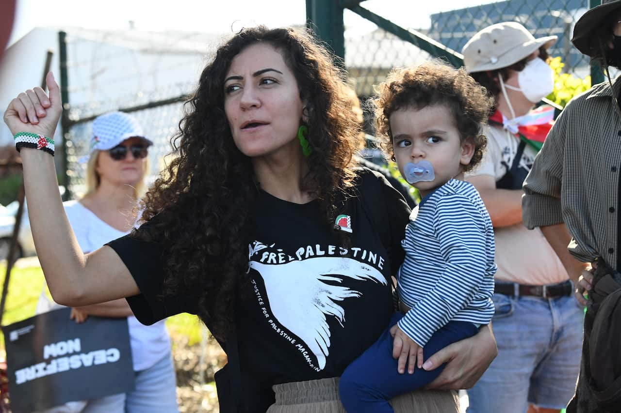 A woman wearing a black T-shirt that reads "Free Palestine" holds a child in her left arm and raises the fist of her other arm. 