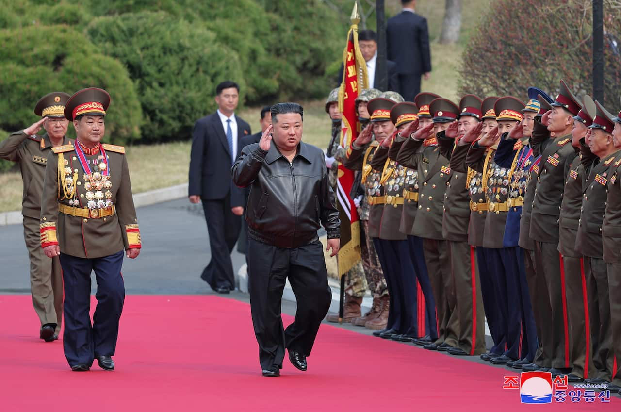 A man in a black leather jacket and dark trousers salutes as he walks down a red carpet past saluting soldiers in uniform