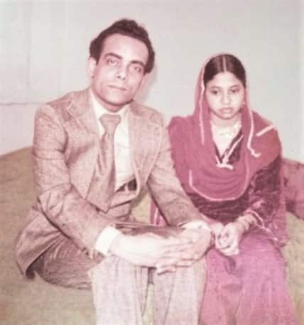 A man in a suit and a woman in a pink shawl sit on a bed.