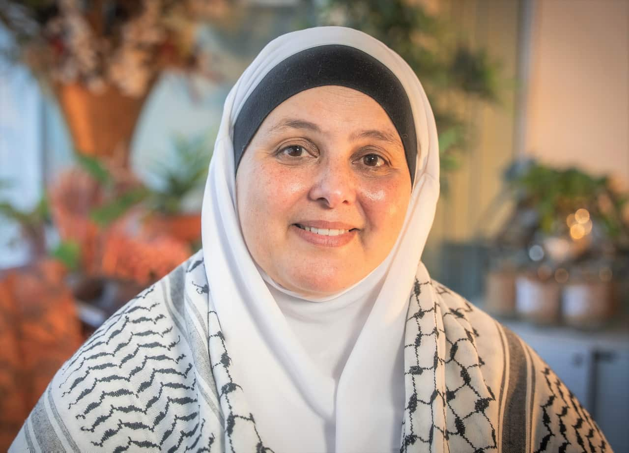 A woman in a shawl smiling at camera and sitting in front of dried flowers.