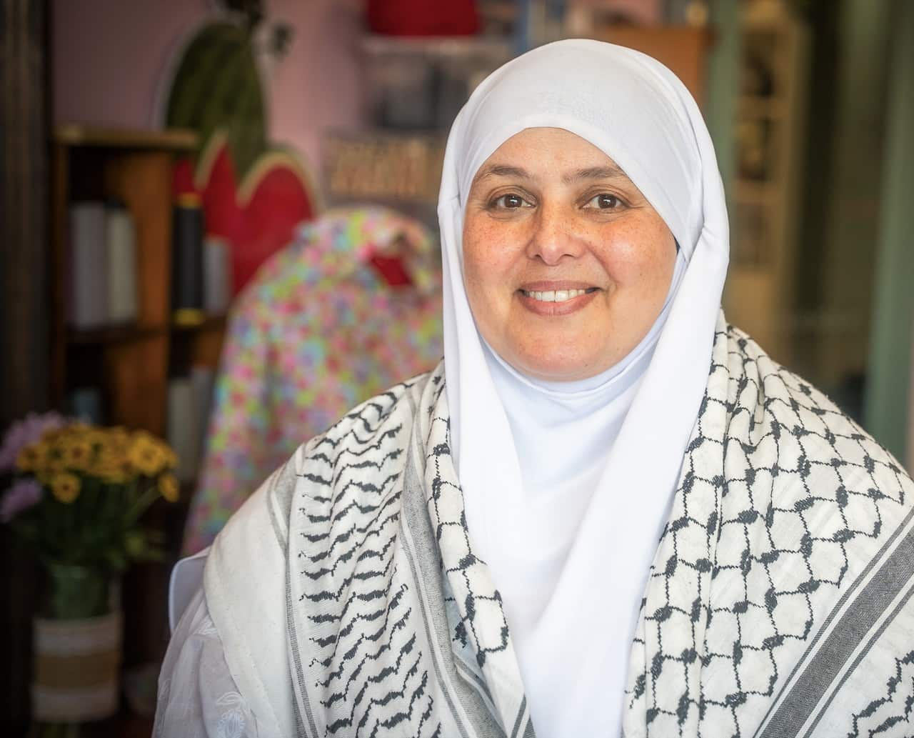 A woman in a shawl, sitting in a sewing workshop with flowers in the background. 