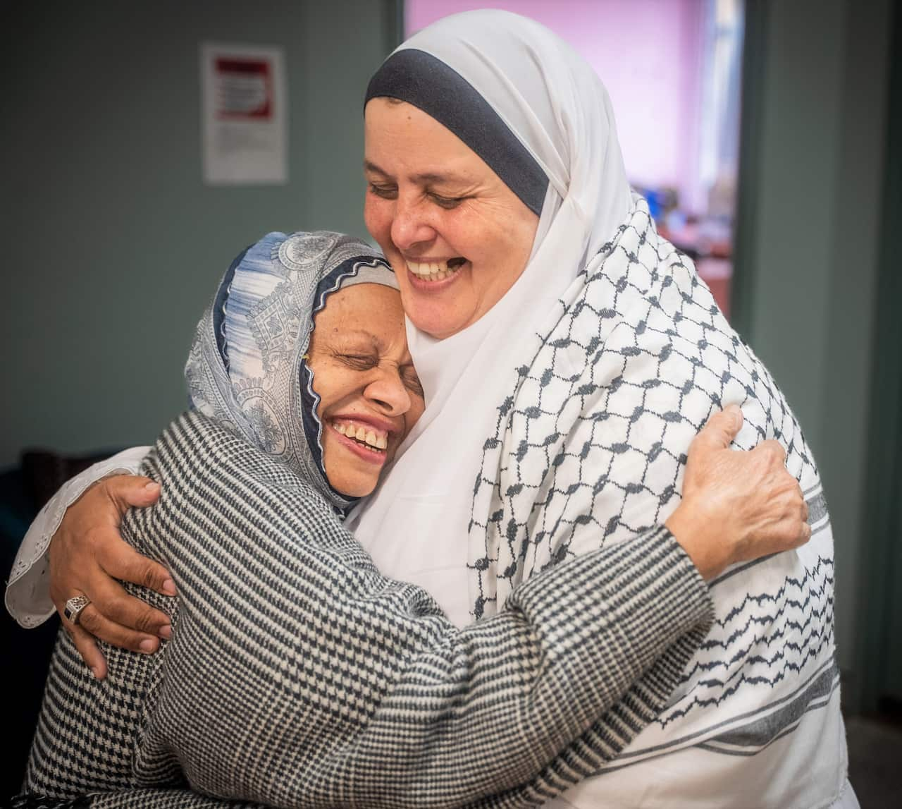 Two women in shawls hug, standing in an office.