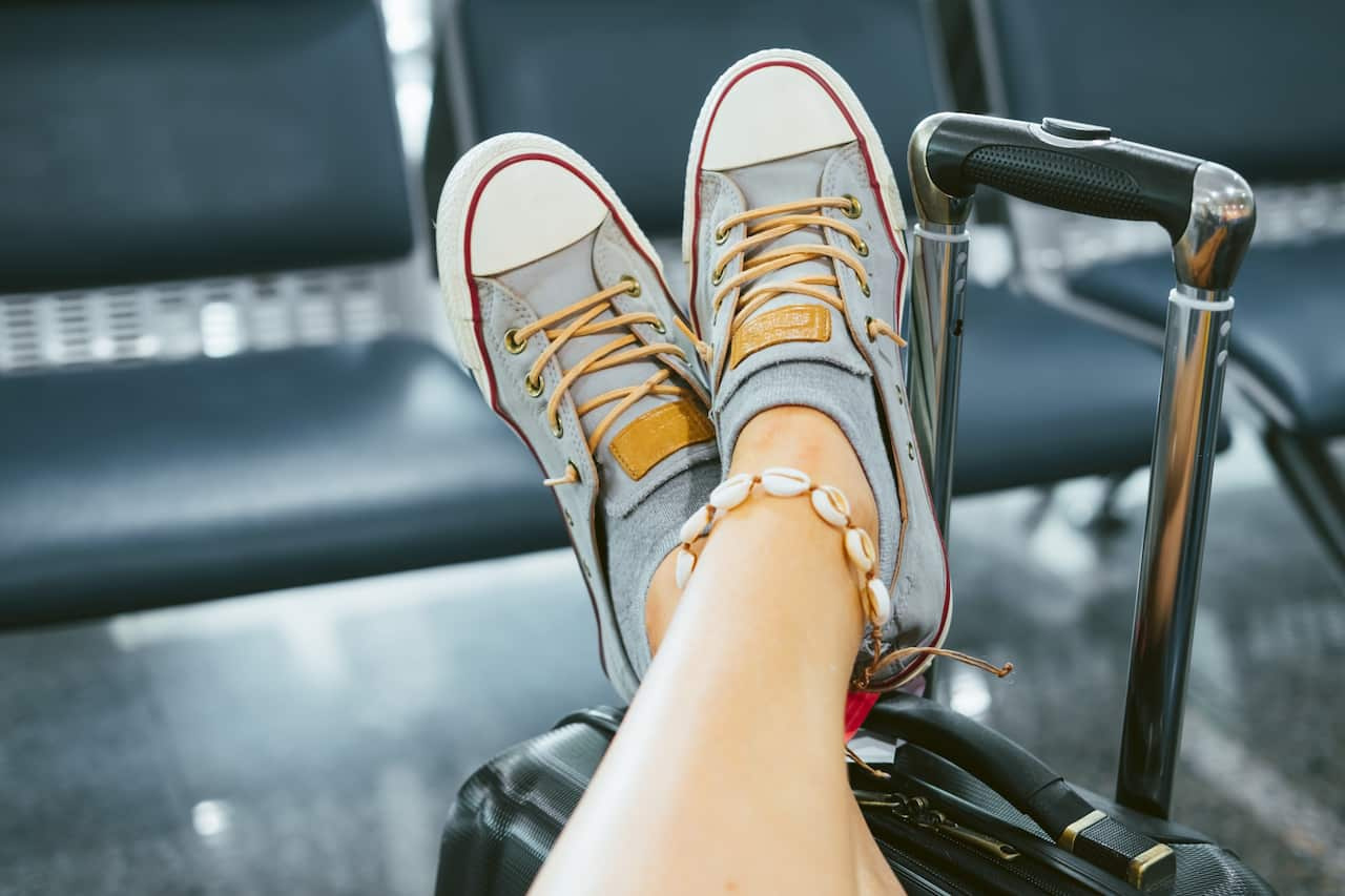 A woman leans her feet on a suitcase. She's wearing blue sneakers and has a shell anklet around one of her ankles.