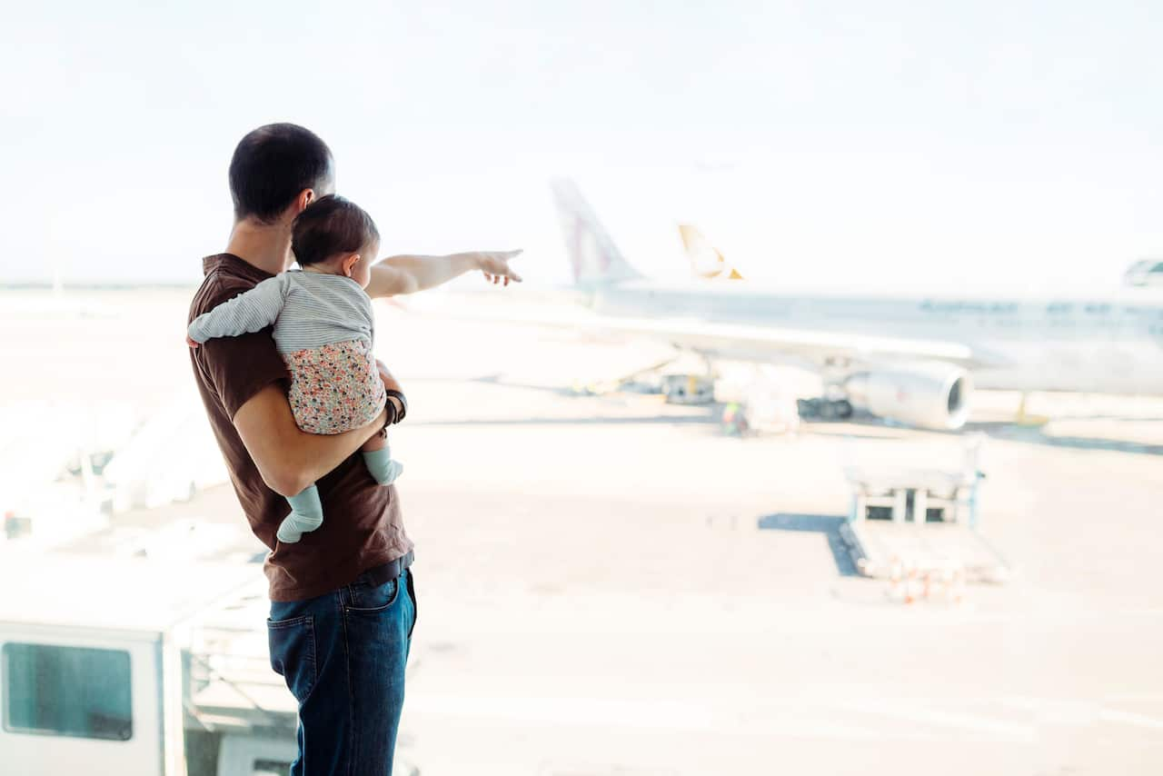 A man holding a baby girl at the airport pointing at the aeroplanes.