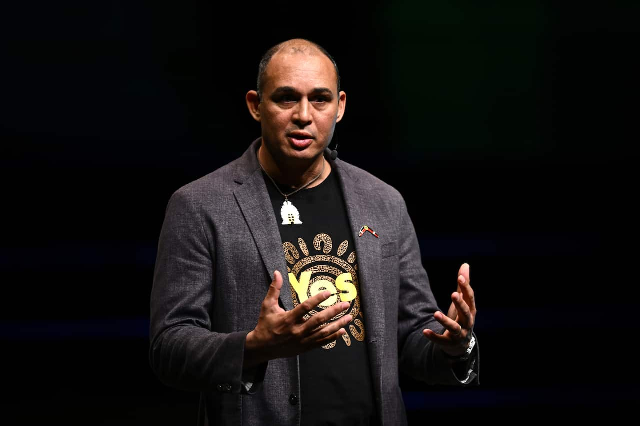 A man in a grey suit jacket and a black t-shirt with Yes written in yellow in the front holds his hands in front of him as he talks