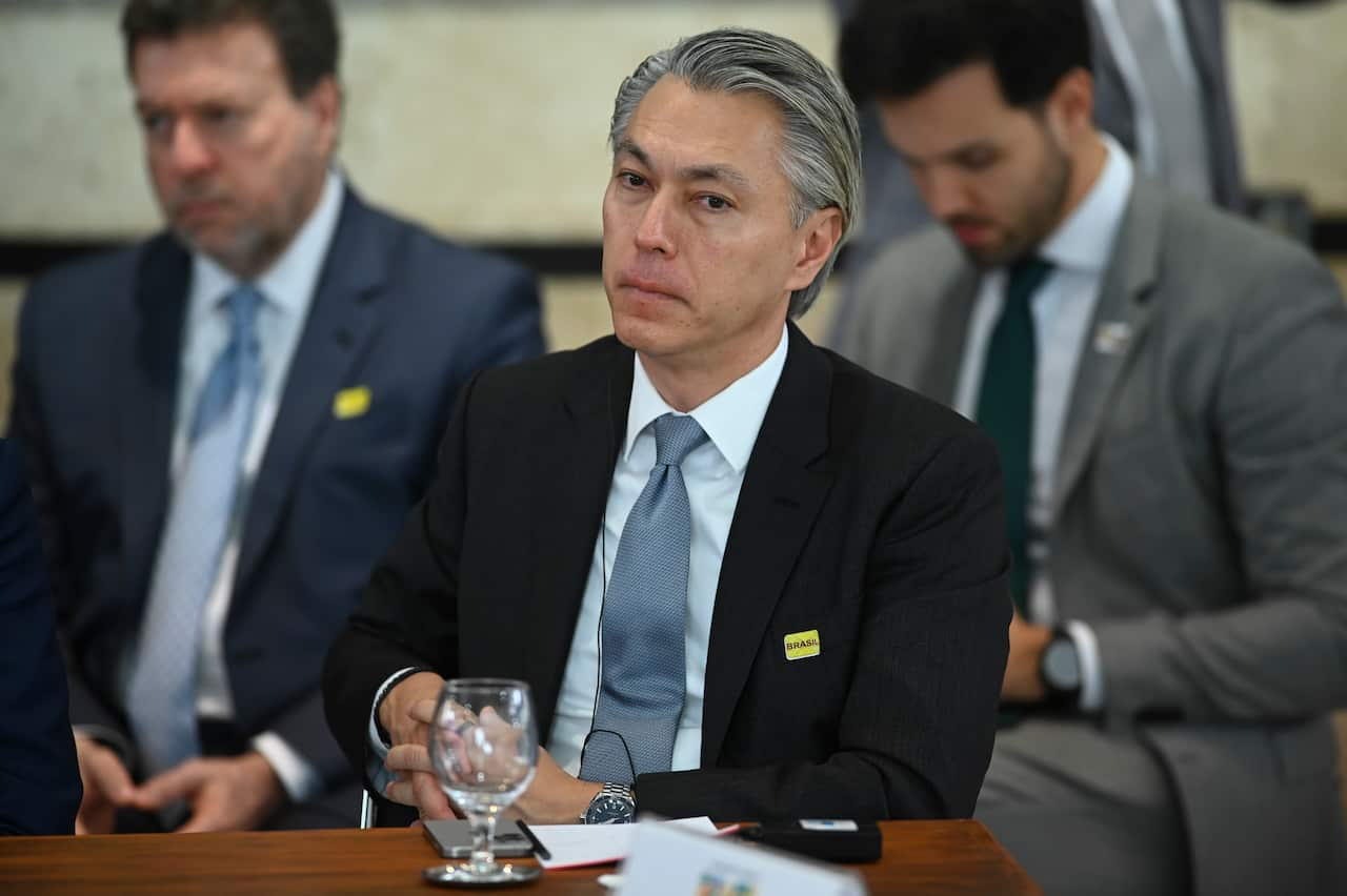 A man in a dark blazer, light blue tie and white shirt sits at a table with a stern expression.