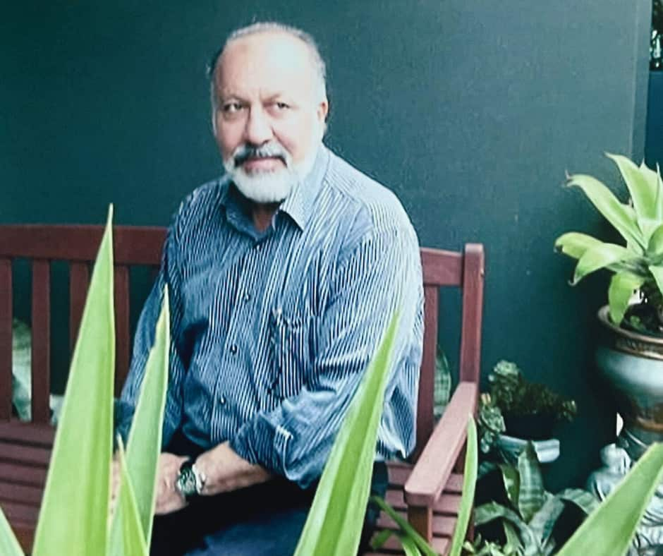 A man with a white beard and wearing a striped shirt sits on a wooden bench, gazing to the side.