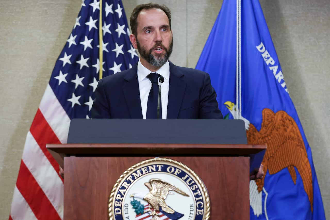 A man in a suit stands at a podium with the US flag and the Department of Justice flag behind him. 