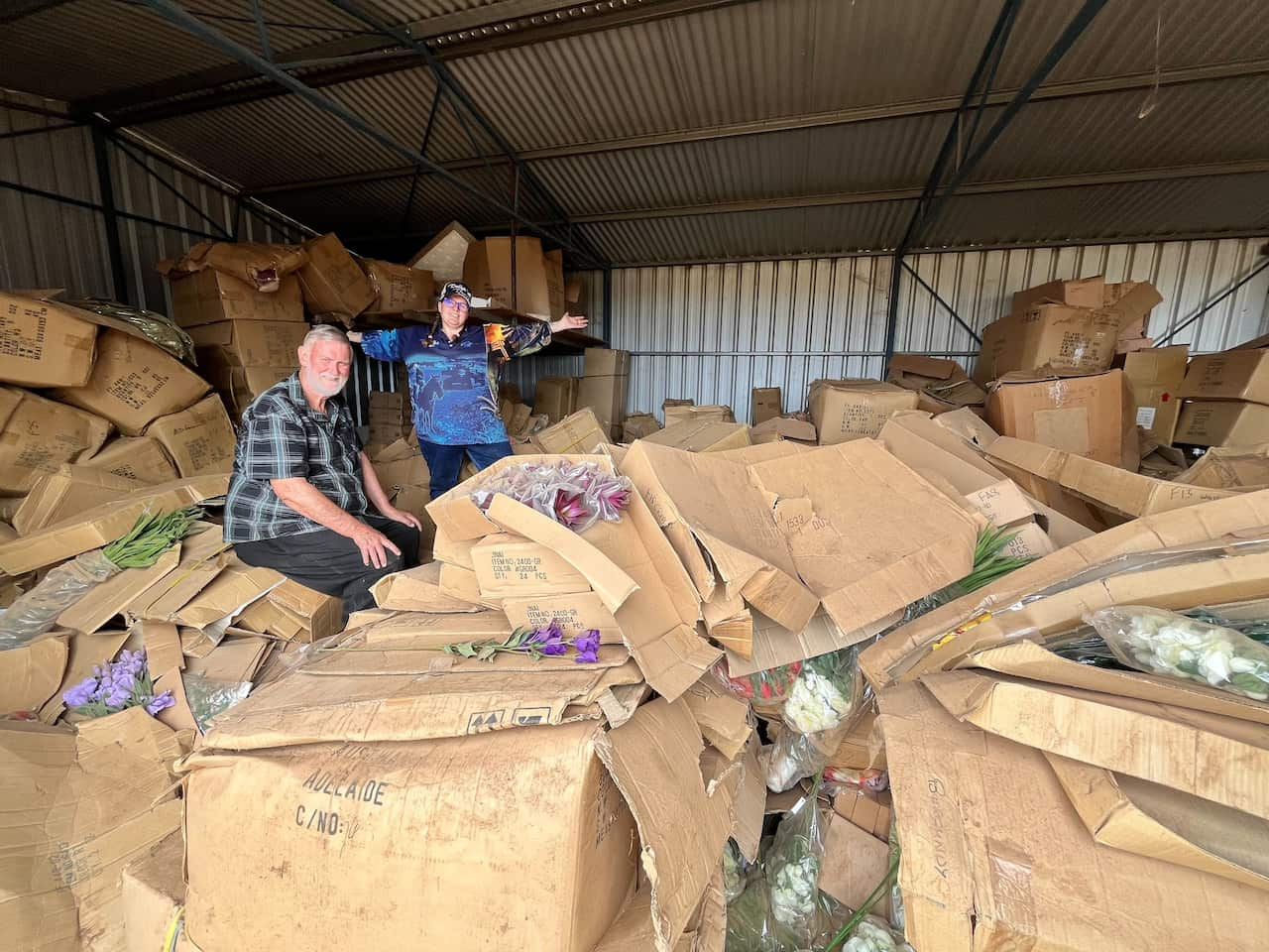 A man and his wife in a shed among boxes of artificial flowers.