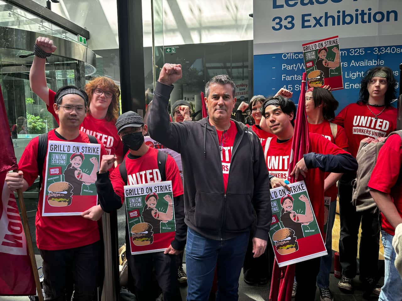 Several people wearing red T-shirts emblazoned with the words "United Workers Union" pose for a photo with their fists raised in the air. In the middle of the photo is an older man wearing a dark grey jacket (union secretary Tim Kennedy). 