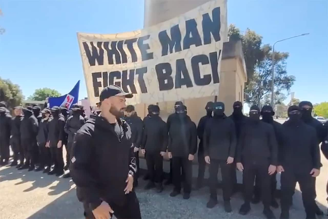 A group of men in black uniforms and balaclavas standing in a row, holding a sign that reads "White Man Fight Back".