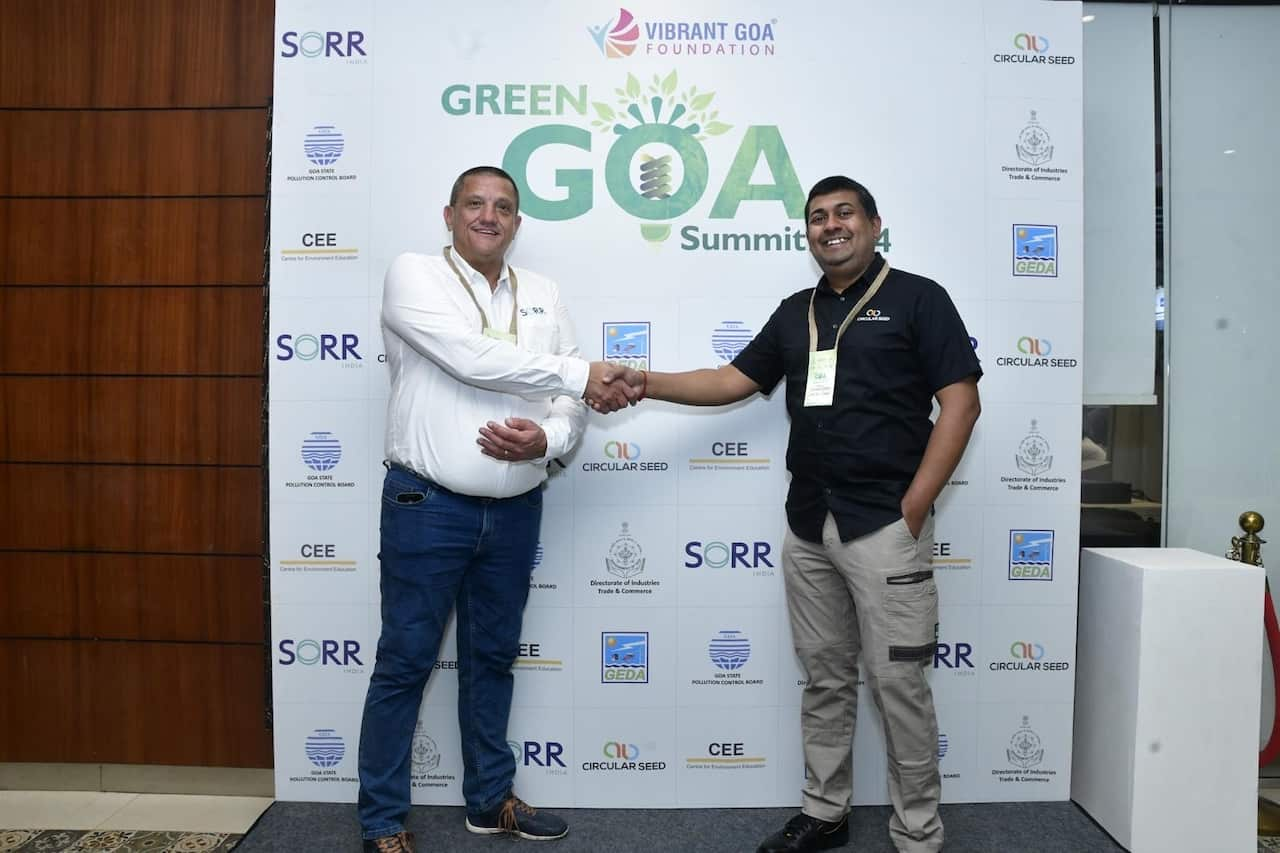 Two men are smiling and shaking hands in front of a poster featuring the words Green Goa Summit and several other logos.