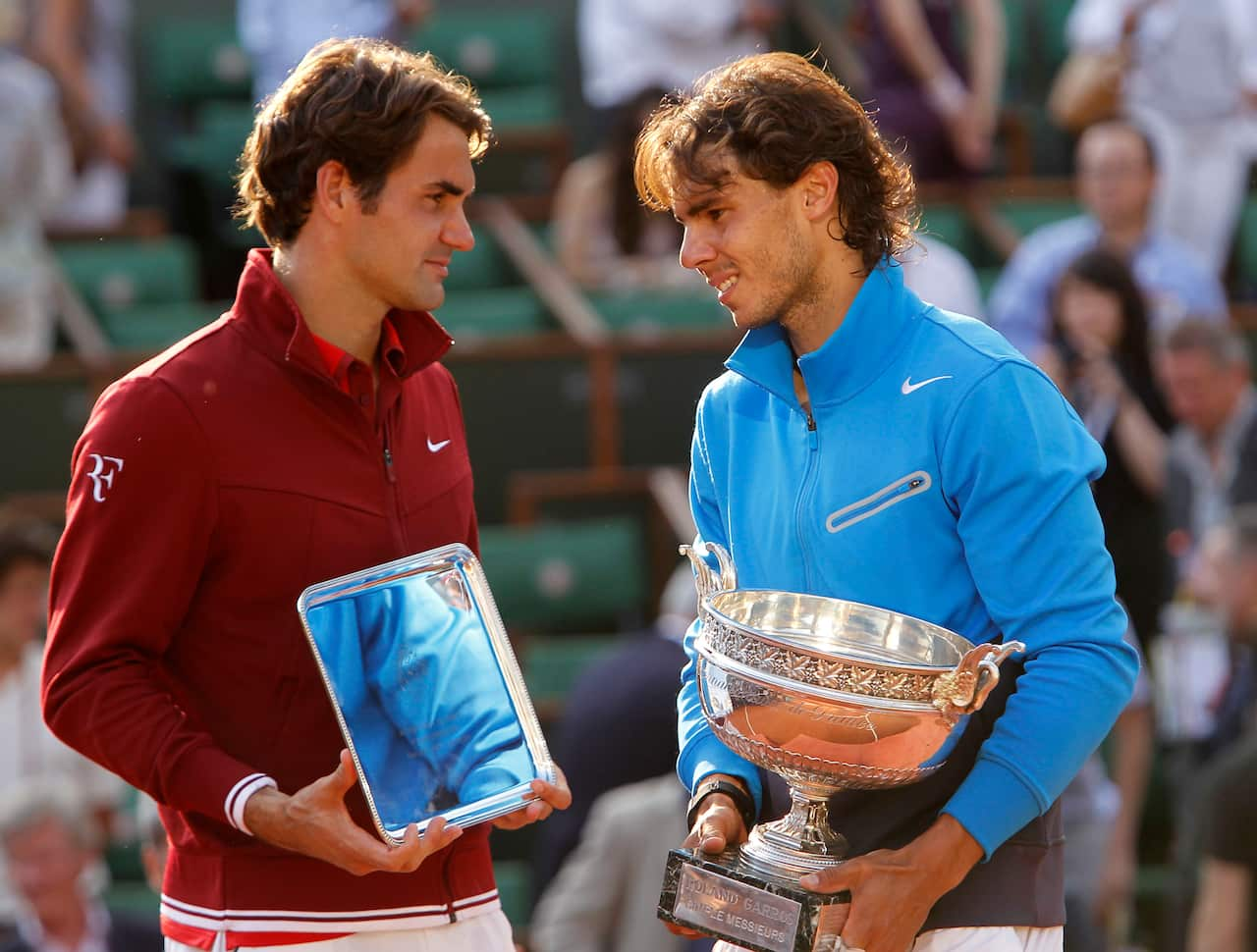 Two men stand facing each other, the one on the left is in a red jacket holding a small box and the one on the right is in a blue jacket holding a large silver trophy 