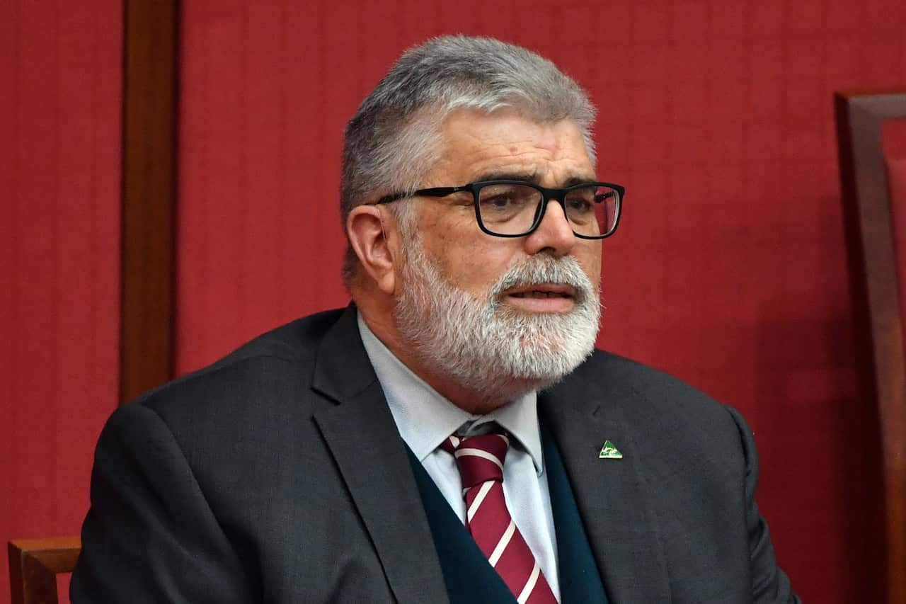 A man in a black suit and a red tie with white stripes sits against a red background.