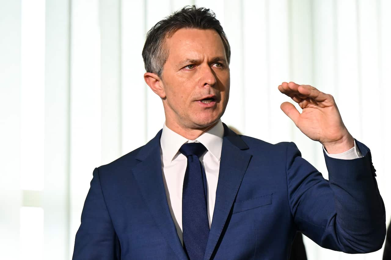 A man in a blue suit and white shirt gestures with his hand while speaking.