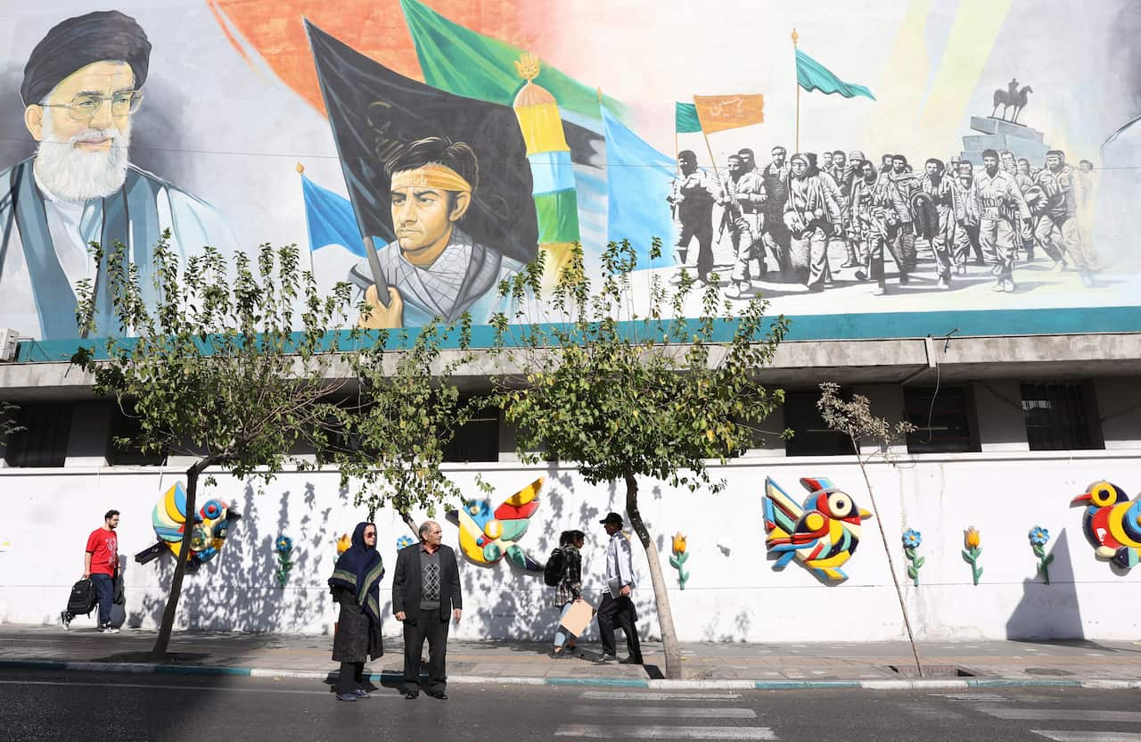 People walking alongside a sidewalk. A large mural above them contains a painting of Iranian Supreme Leader Ayatollah Ali Khamenei.