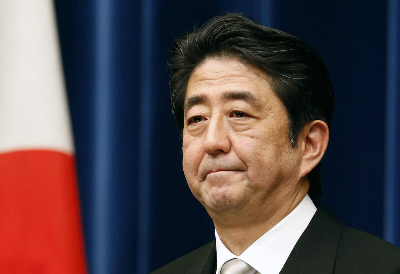 Shinzo Abe standing in front of a blue curtain. He is wearing a black suit and white shirt.