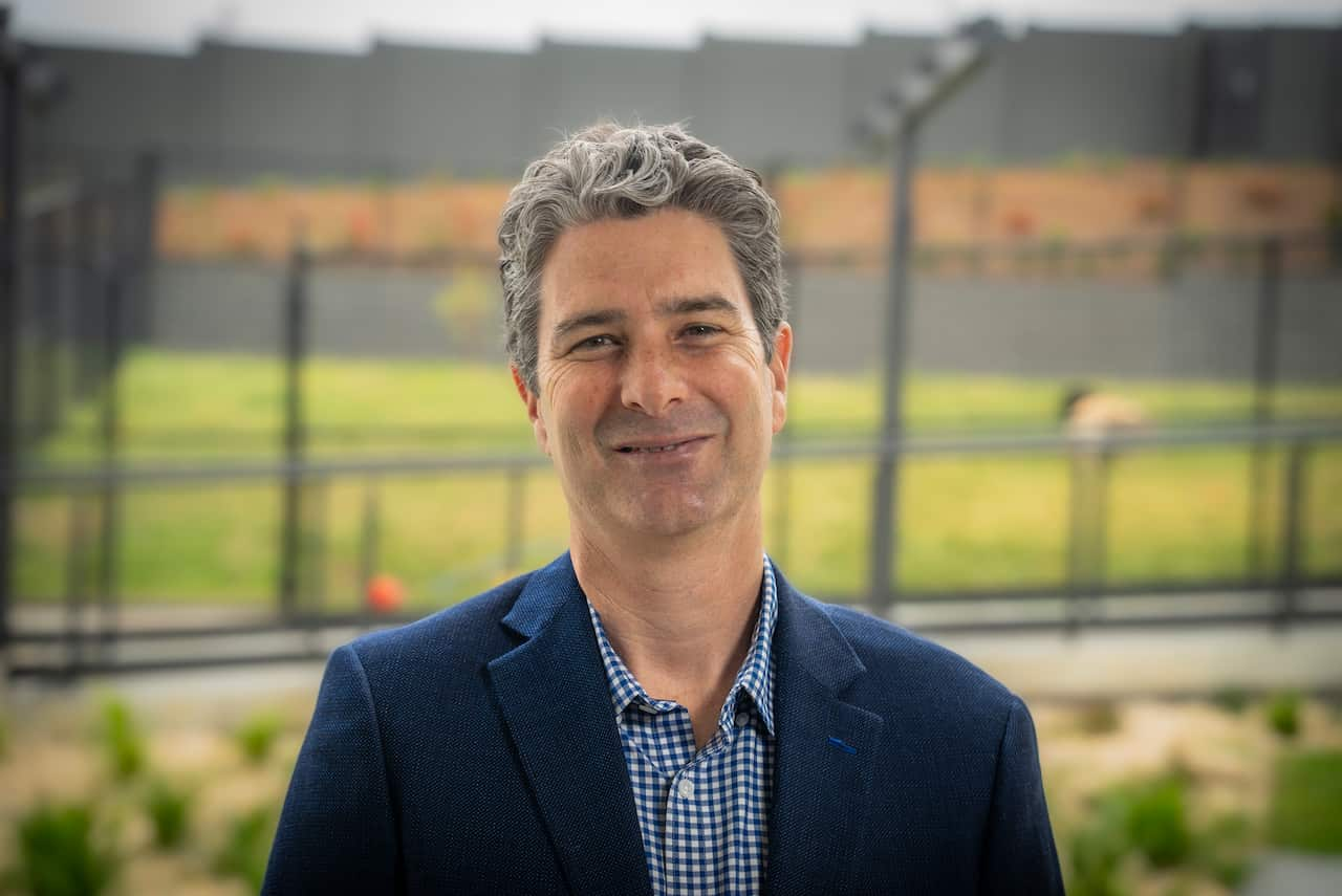 A man in a blue blazer is smiling at the camera in front of a fenced yard.