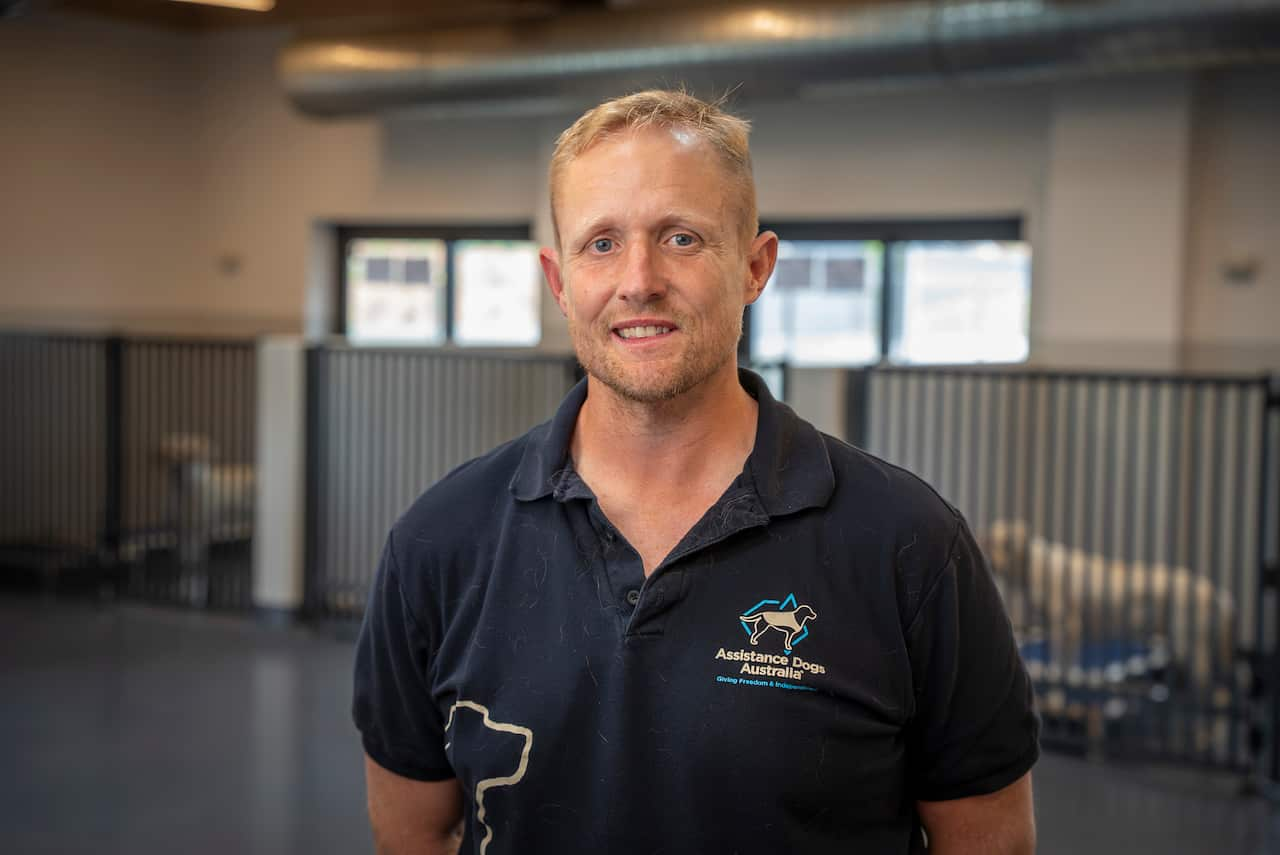 A man wearing a t-shirt featuring the Assistance Dogs Australia logo is smiling at the camera. Behind him, there are dogs in kennels.