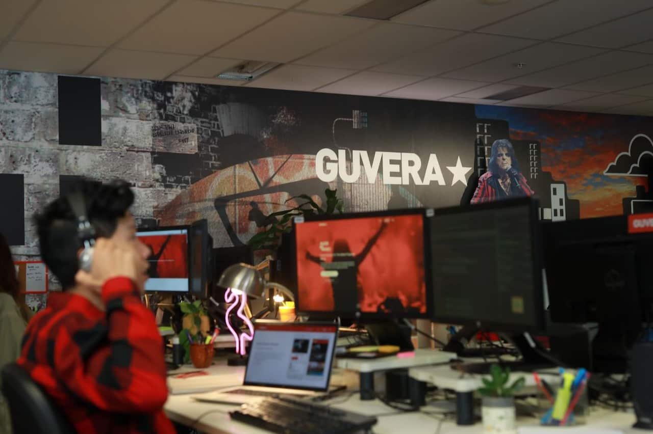 A person wearing headphones and a red chequered shirt sits at a desk looking at two computer screens. Behind is a mural with the word Guvera written on it and a cardboard cutout of Alice Cooper