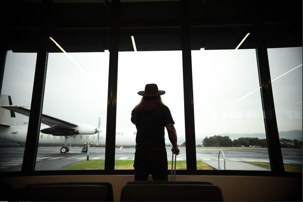 A man with heavily tattooed arms and a wide-brimmed hat stands silhouetted against a wide glass window, looking outside towards a plane on the tarmac 