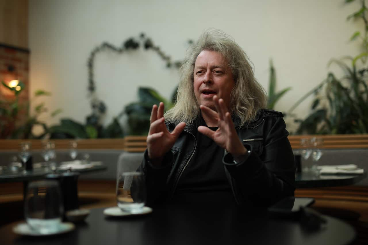 A man with shoulder-length grey hair and a leather jacket sits in a restaurant. He gestures in an explanatory manner front of his face