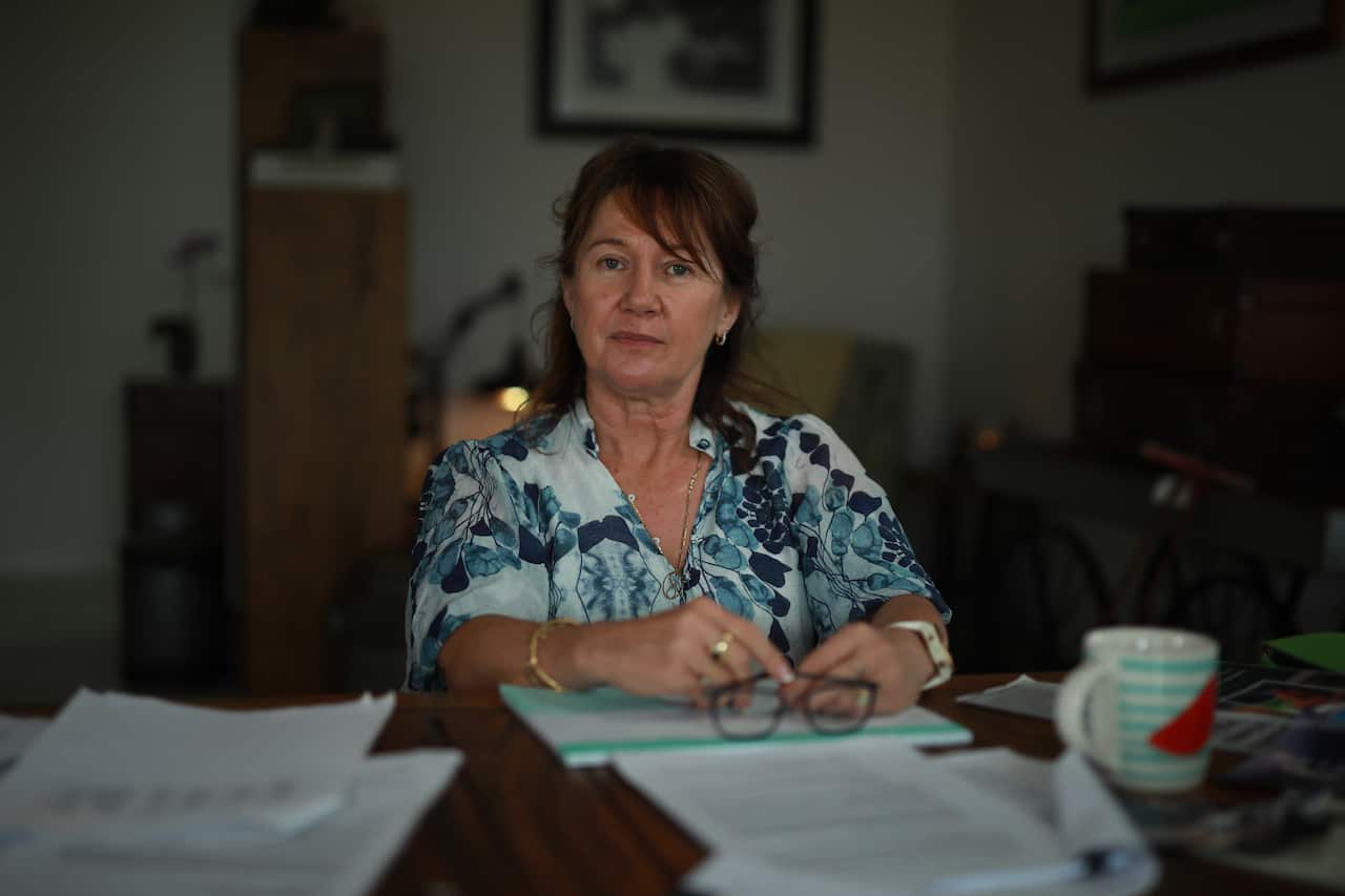 A woman in a blue and white floral top stares solemnly at the camera while holding her glasses atop a desk covered in loose papers