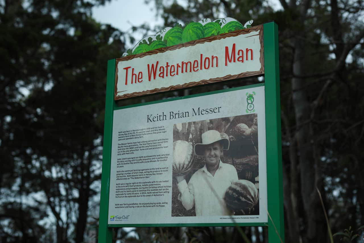 A green sign board stands amongst trees, titled The Watermelon Man. It features a black-and-white photo of a man holding two watermelons and a biography of Keith Brian Messer