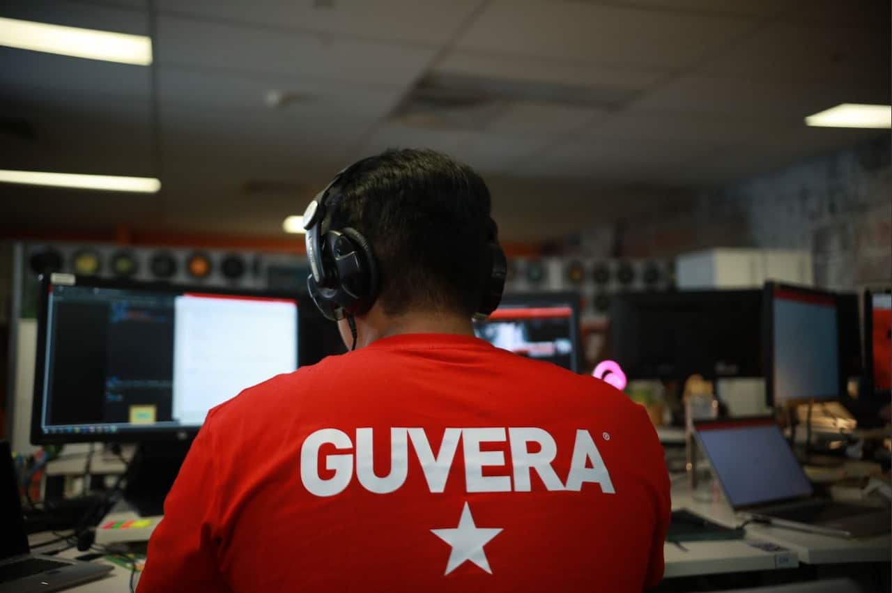 A man at a desk with his back to the camera, surrounded by multiple computer screens. He is wearing a red T-shirt with the word Guvera and a white star on the back