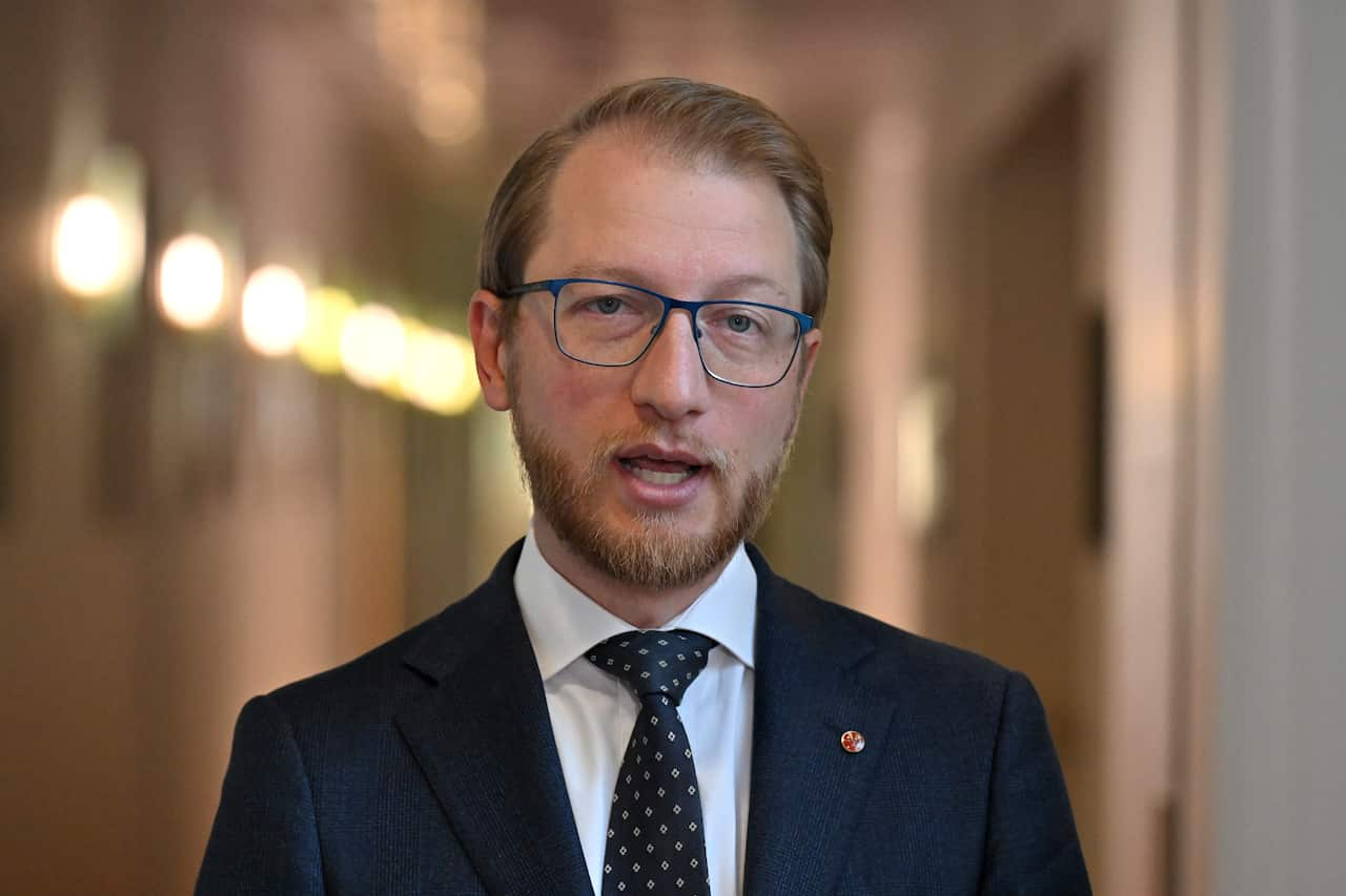 James Paterson, wearing blue glasses, wearing a suit and standing in a hallway.