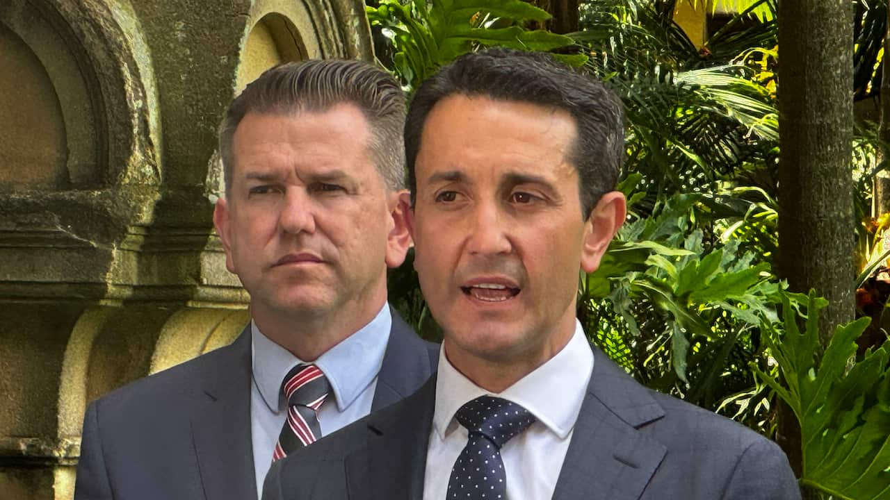 A man in a grey-blue suit speaking in front of bushes. Another man is behind him.