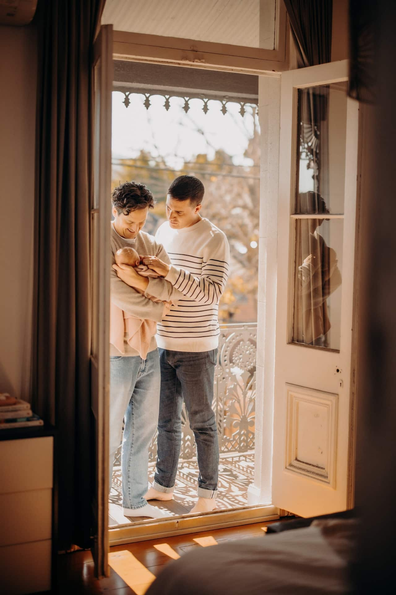 Two men stand on a balcony holding their baby. 