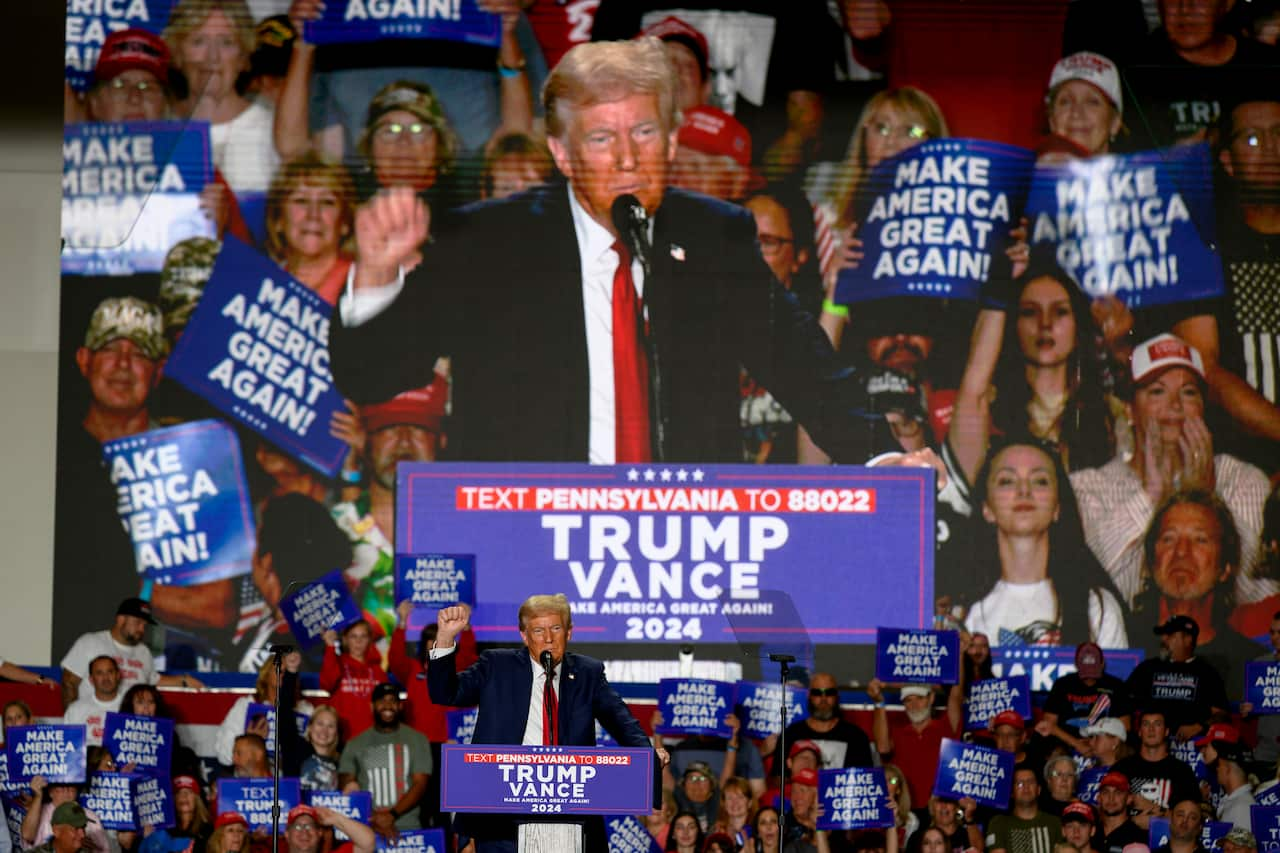 Donald Trump stands in front of a podium with 'Trump Vance' signs in the background.