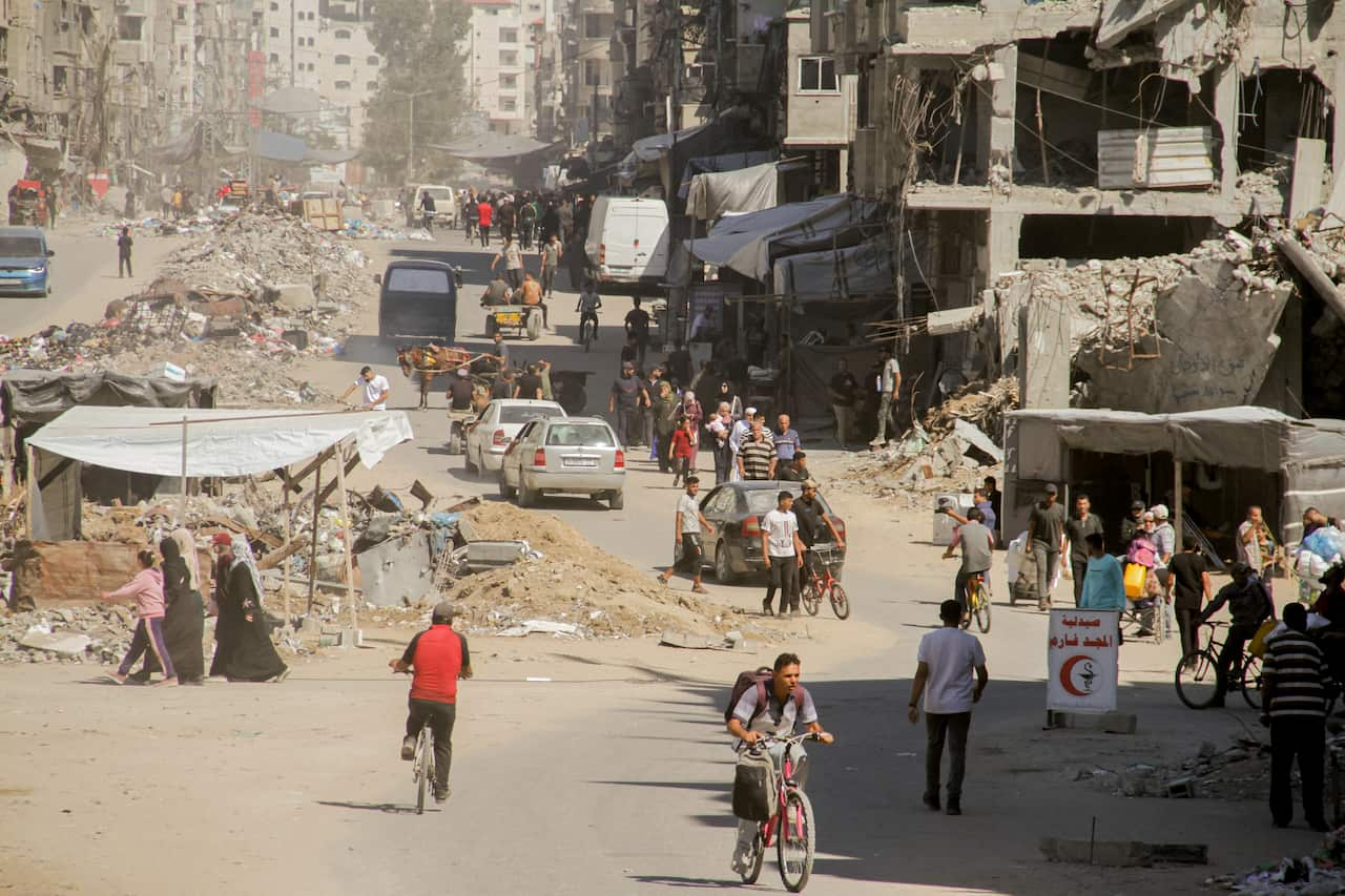 People are walking and others are cycling among the ruins and debris of a city. Some vehicles can also be seen on the road.