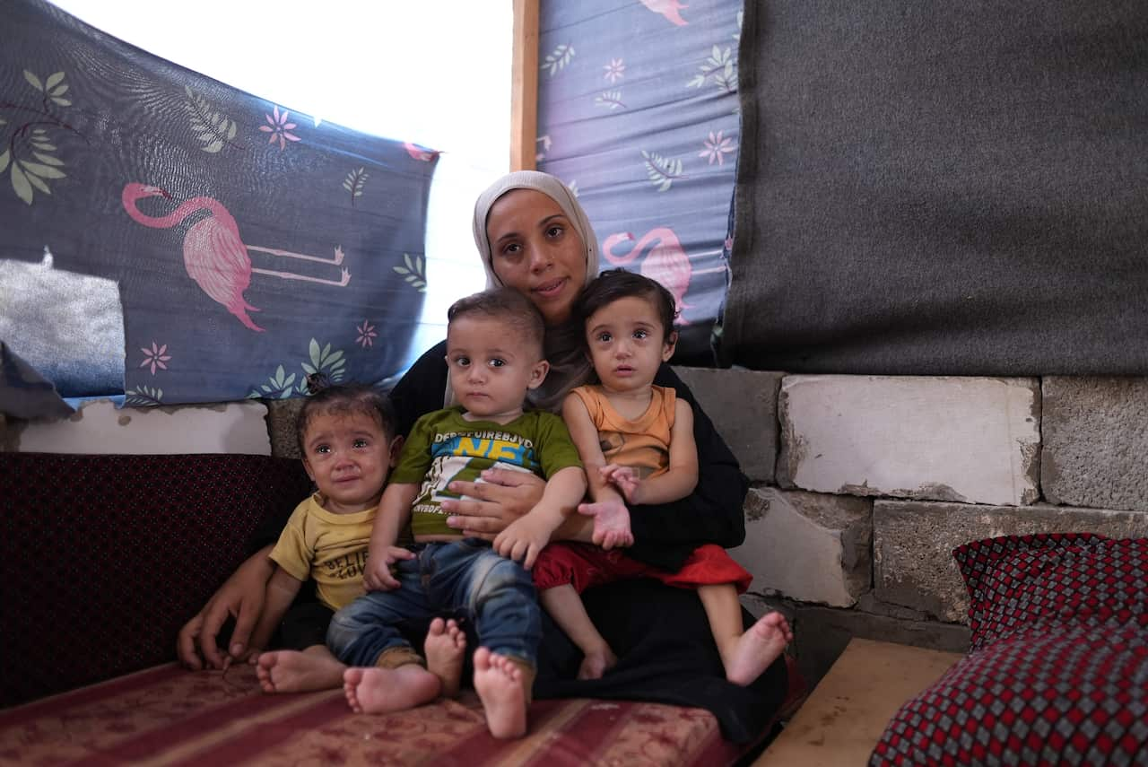 A woman sits in a makeshift living room with three small children in her arms. 