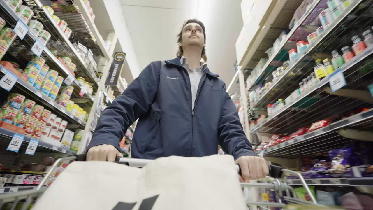 A man in a blue jacket pushes a trolley through a supermarket aisle 