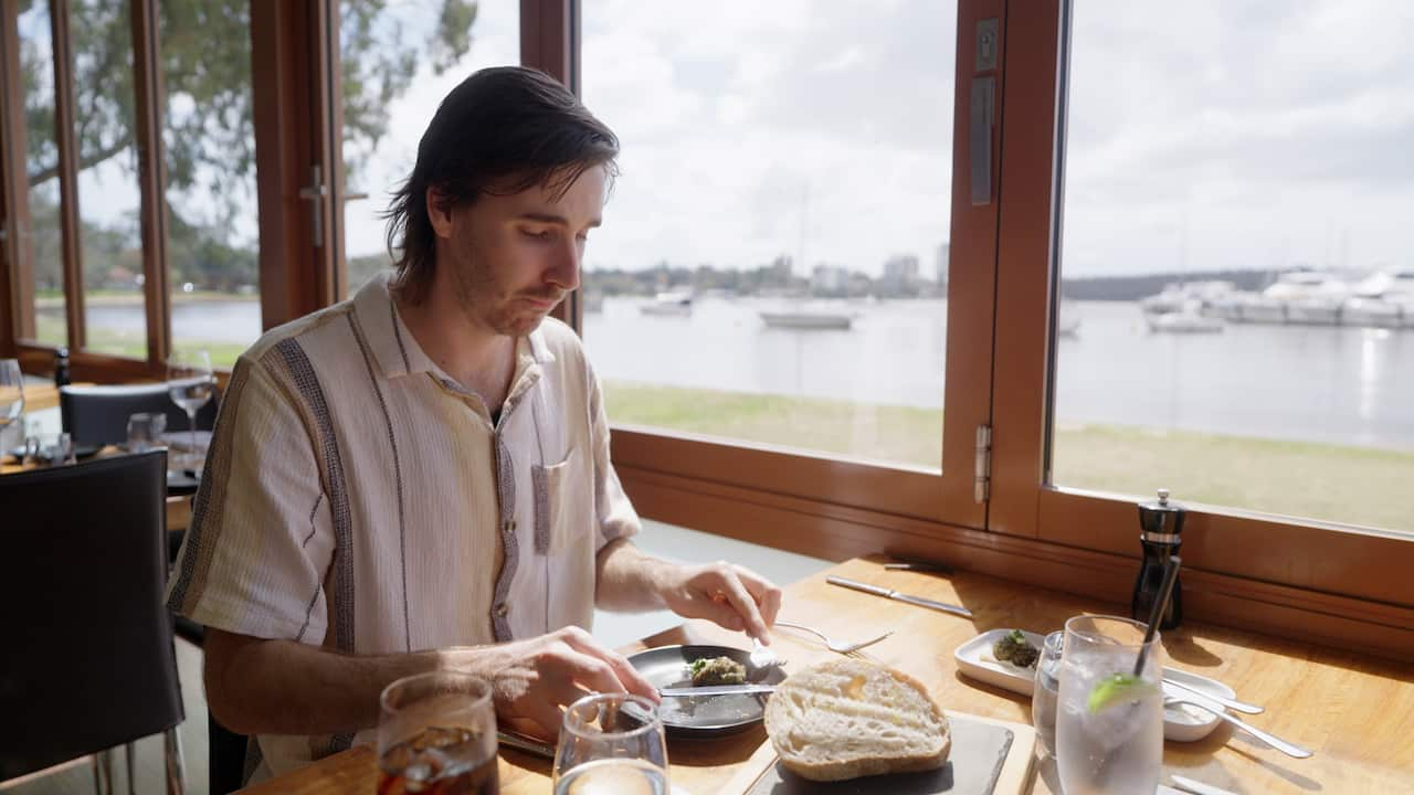 A man in a striped shirt sits in a waterfront restaurant, making a disgusted face while looking down at a zucchini fritter on his plate