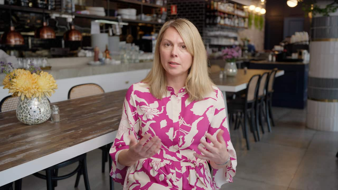 A blonde woman in a pink floral dress looks at the camera while gesturing in an explanatory manner with her hands