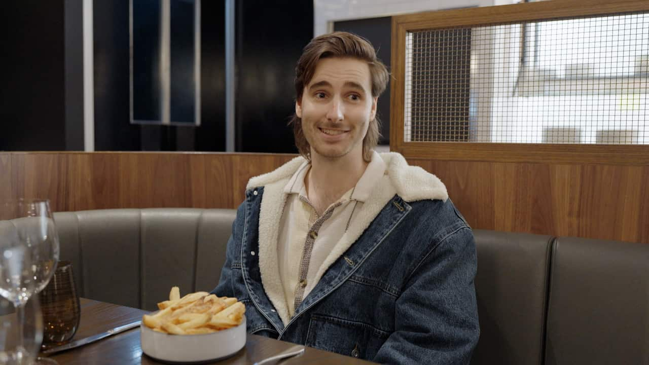 A man in a denim jacket smiles while sitting in a restauran with a bowl of chips in front of him.