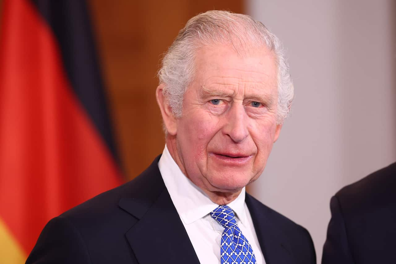 A man (King Charles) with white hair in a dark suit looks slightly to his right with a serious expression on his face. 