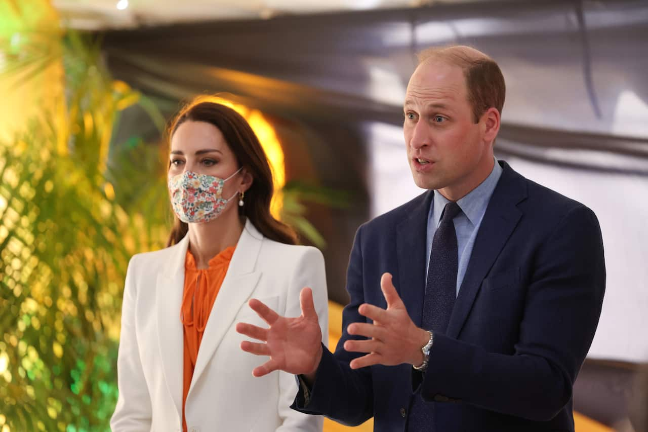 A woman wearing a mask and a white jacket stands next to a man in suit gesturing as he speaks.  