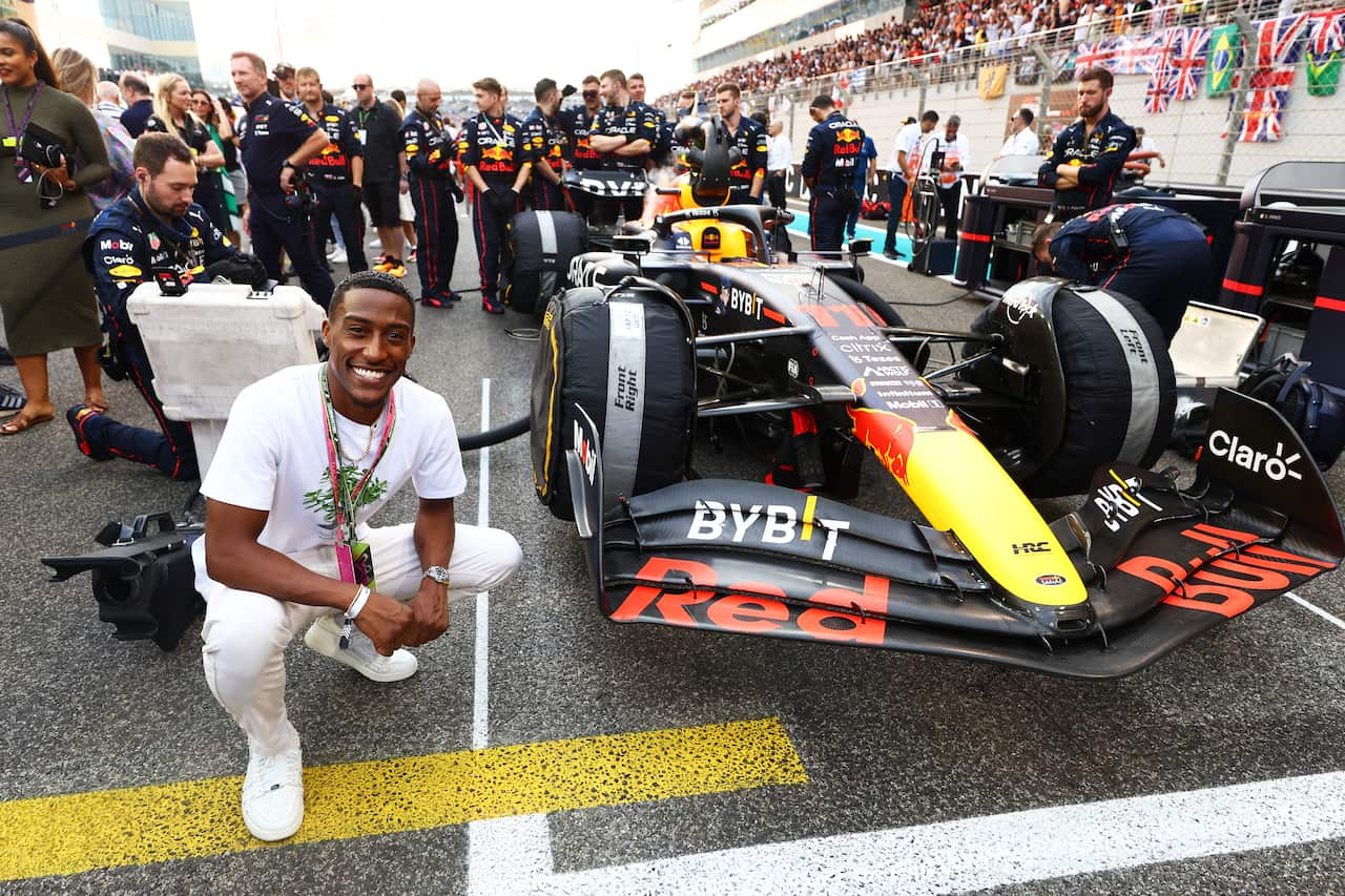 A man wearing a white t-shirt and pants is posing next to an F1 vehicle on the grid ahead of racing.