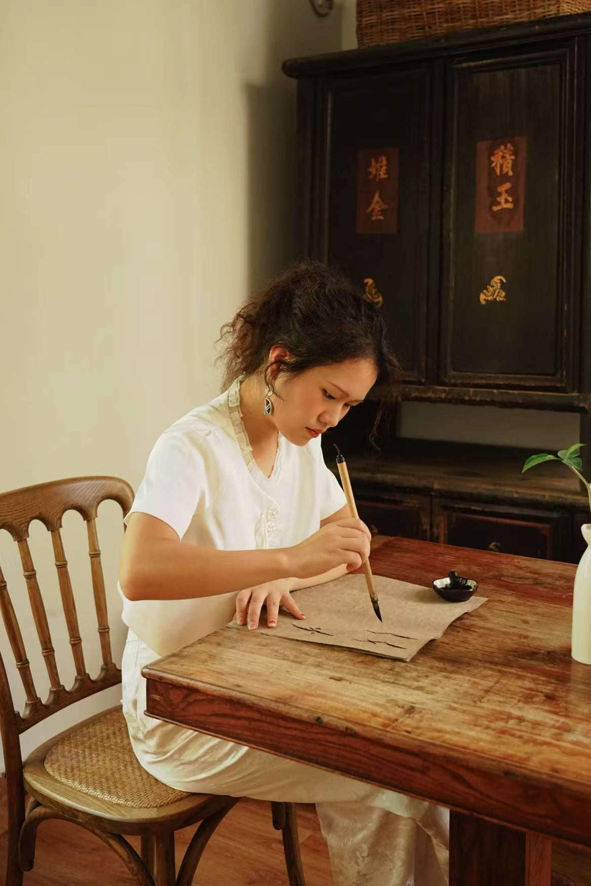 A woman in a white shirt holds a long brush and paints onto brown paper at a wooden desk.