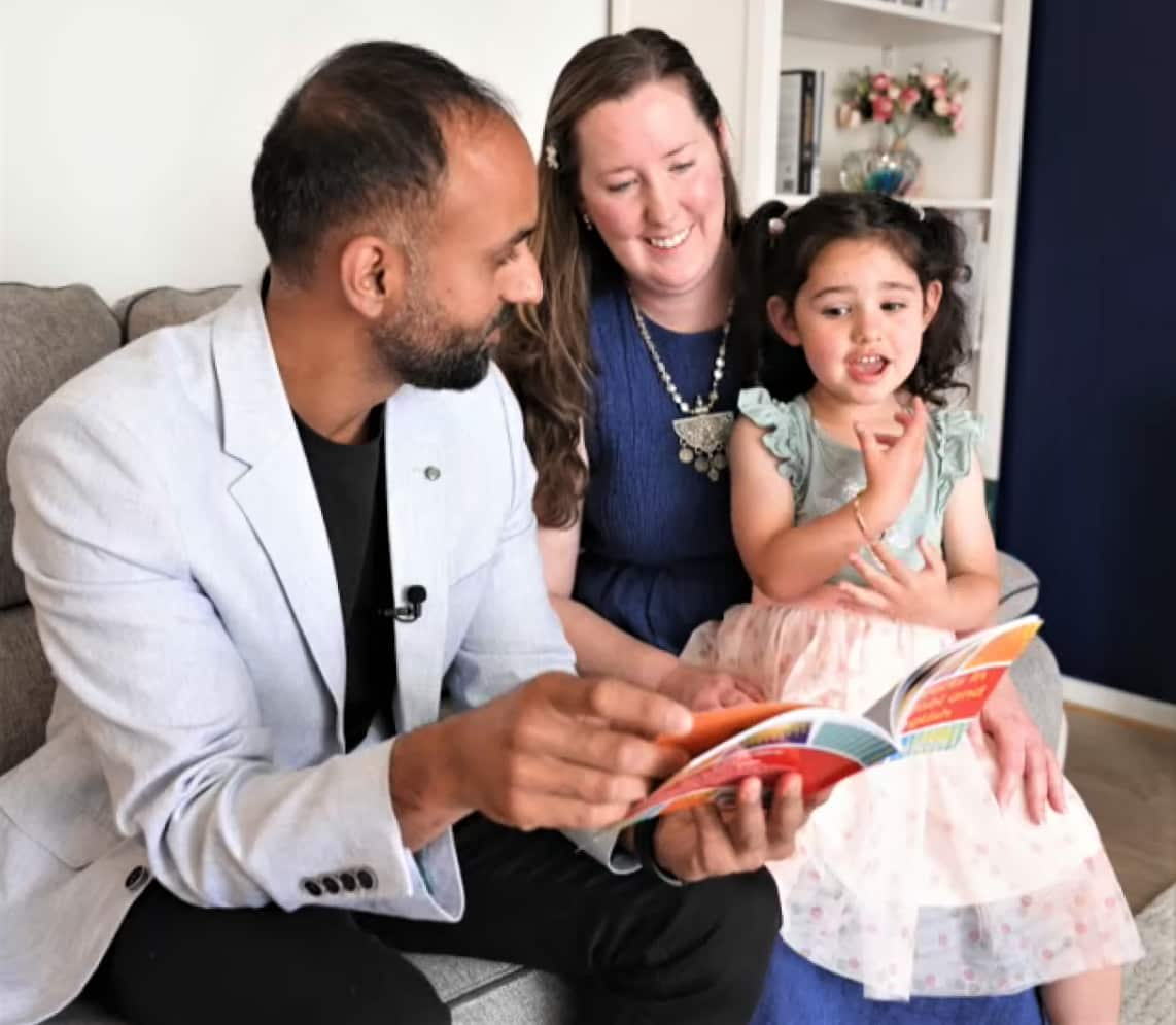 A man holds a book while a woman in a blue dress and a child look on.