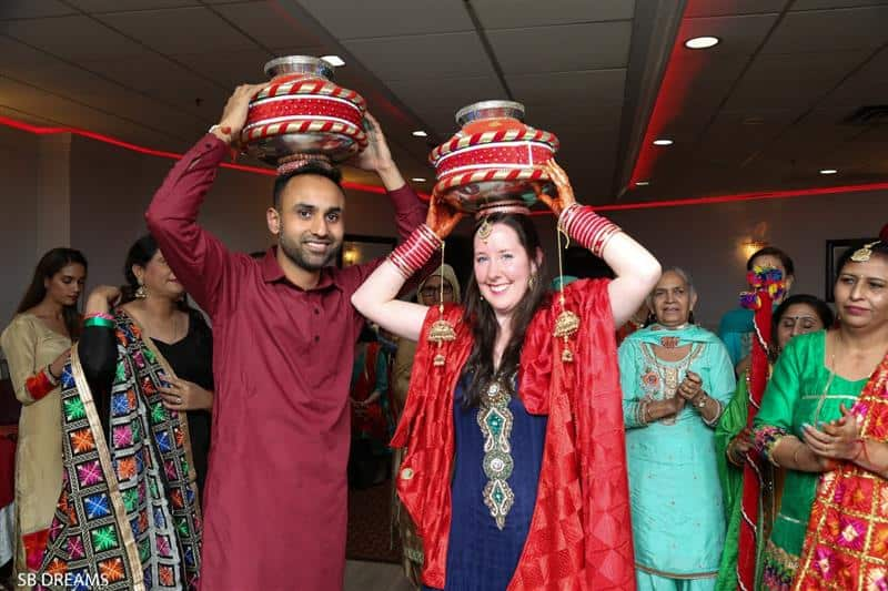 A couple wearing red ceremonial dress smile at the camera holding traditional pots on their heads. People smile and clap behind them.