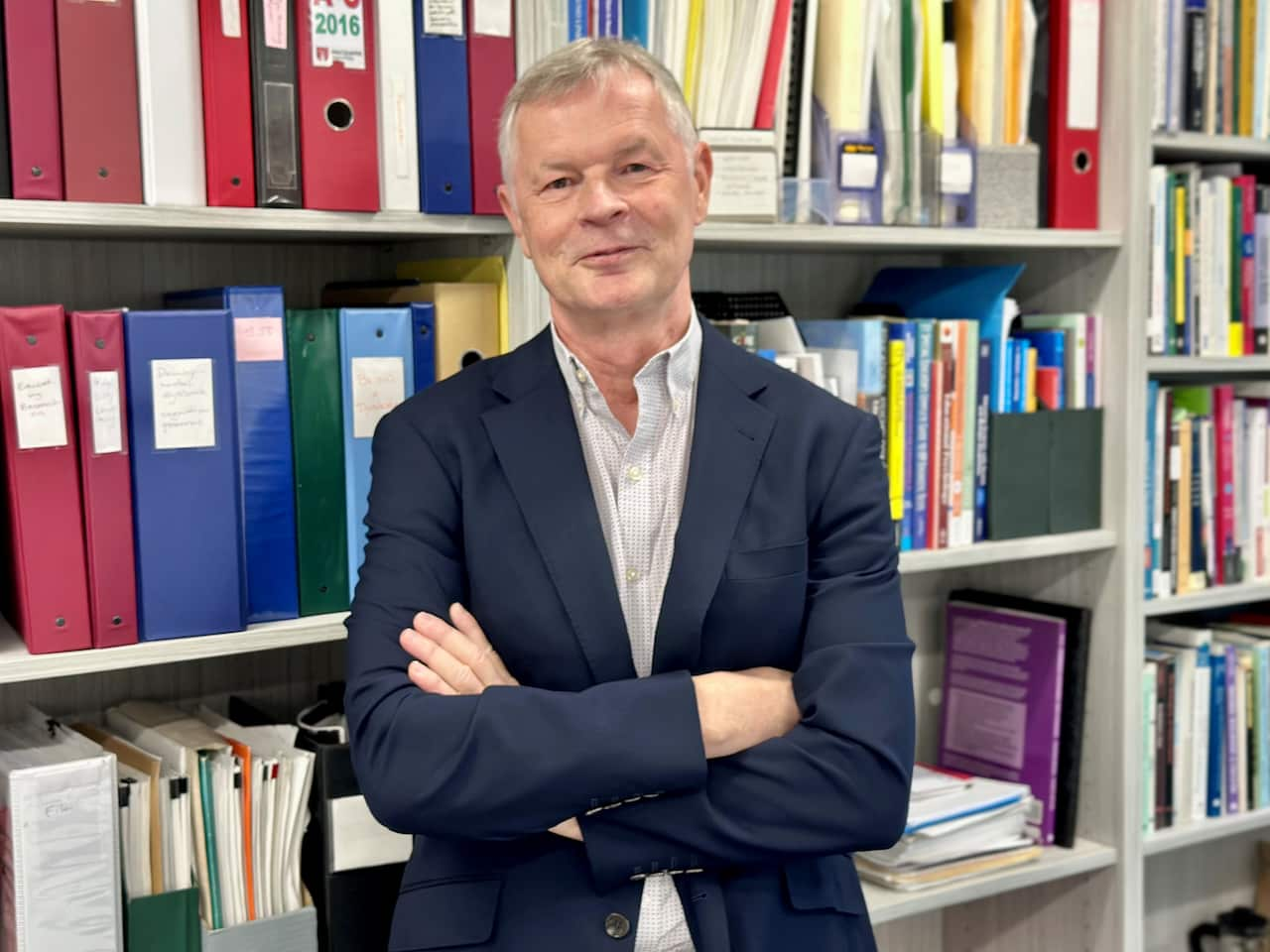 A man smiles to camera with his arms crossed with a colourful bookshelf behind him. 