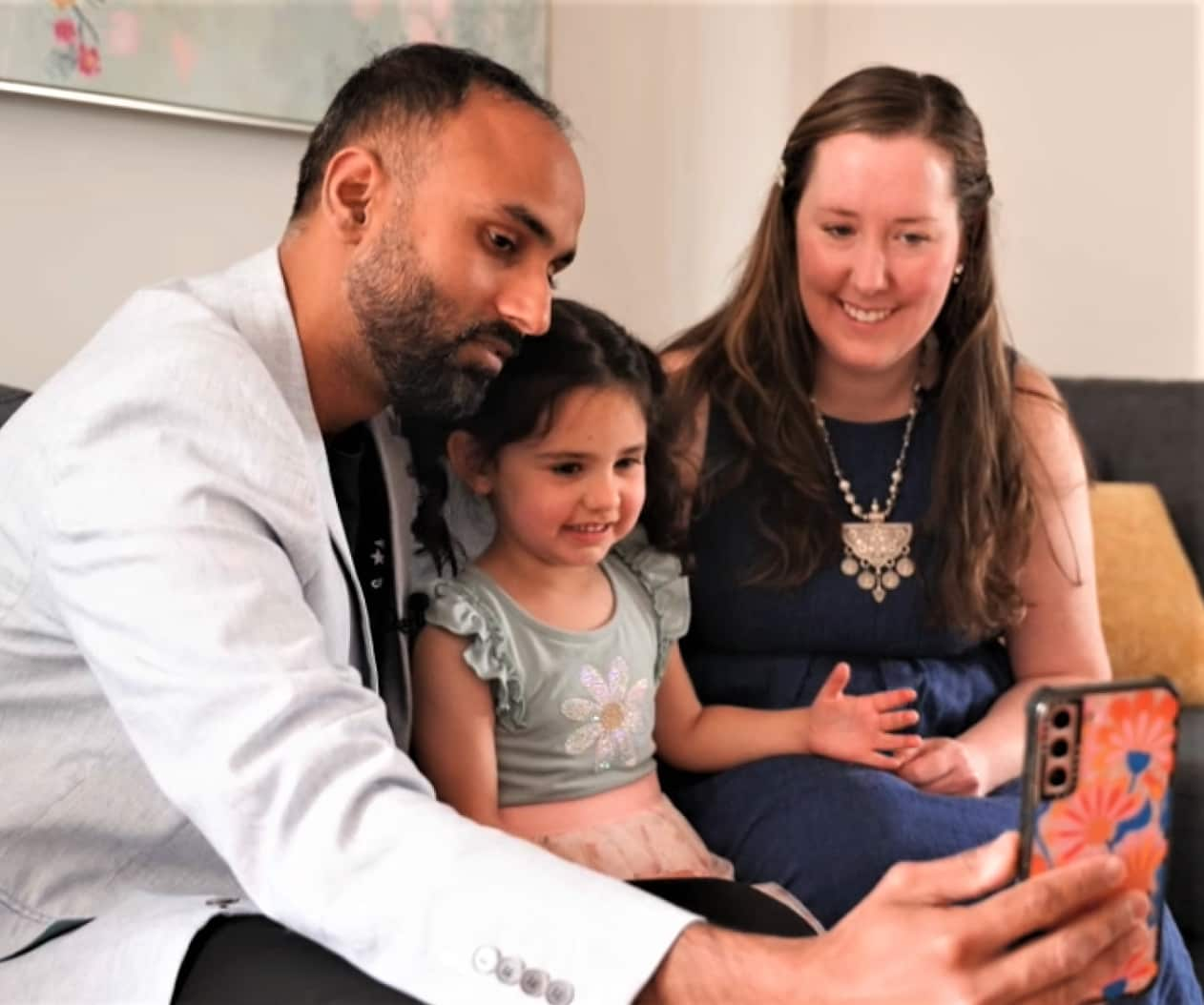 A man in a suit jacket holds a mobile phone while a woman in a blue dress and a child look on. 