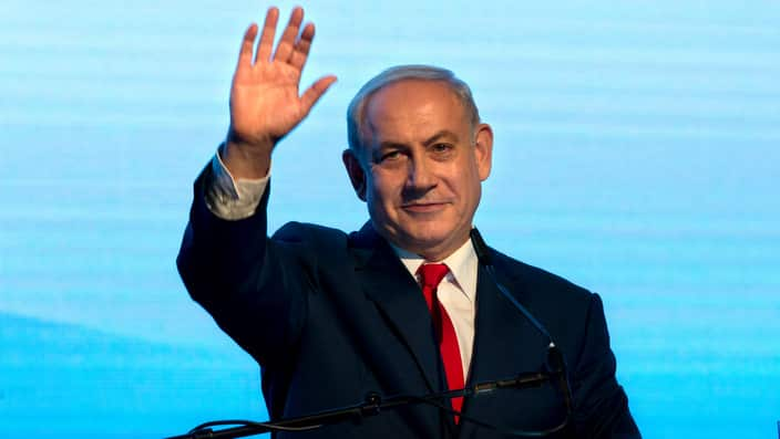 A man in a suit waves in front of a blue screen.