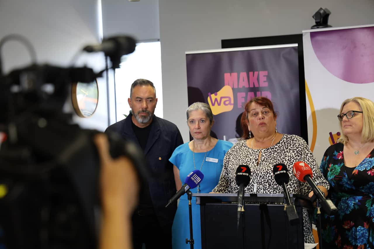 A woman stands behind a lectern with microphones in front of it. There is a woman standing to her left and a woman and a man standing to her right