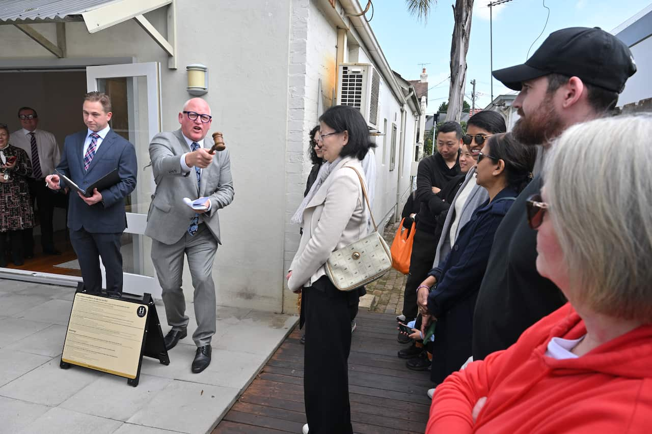 An auctioneer, wearing a grey suit, points his gavel towards a woman in the crowd during an auction.